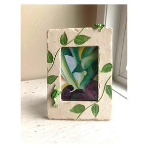 Puede incluir: Marco de fotos rectangular con un acabado texturizado en blanco roto. Hojas verdes y pequeñas figuras de ranas verdes decoran el marco. El marco contiene una imagen abstracta colorida. Un acento decorativo para la decoración del hogar.