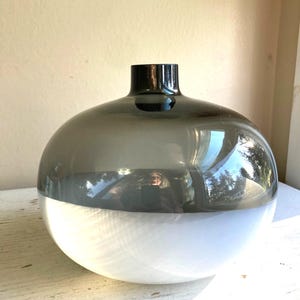 May include: A two-toned glass vase with a rounded body and a short, cylindrical neck. The top half is a smoky gray, transitioning to a clear, white base. The vase reflects the surrounding environment.