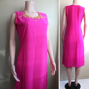May include: A sleeveless pink dress with gold embroidery around the neckline. The dress has a sparkly texture and a straight silhouette.