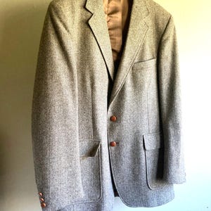 May include: A light brown tweed blazer with a herringbone pattern hangs on a wooden hanger. The blazer has two front pockets, brown buttons, and a brown satin lining. The jacket is a classic style.