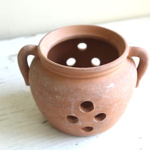 May include: A terracotta pot with two handles and several holes. The pot has a round body and a wide opening. The colour is a warm, earthy tone, suggesting a rustic or handmade aesthetic. The holes are evenly spaced.