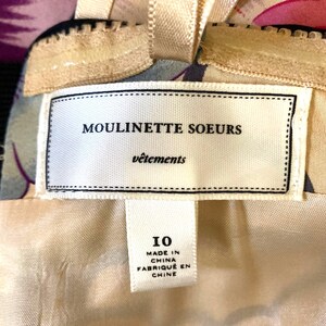 May include: A garment label featuring the text MOULINETTE SOEURS v&ecirc;tements with a size 10 tag below it. The label is white with black text and is attached to a beige fabric with a floral pattern. The tag also states MADE IN CHINA.