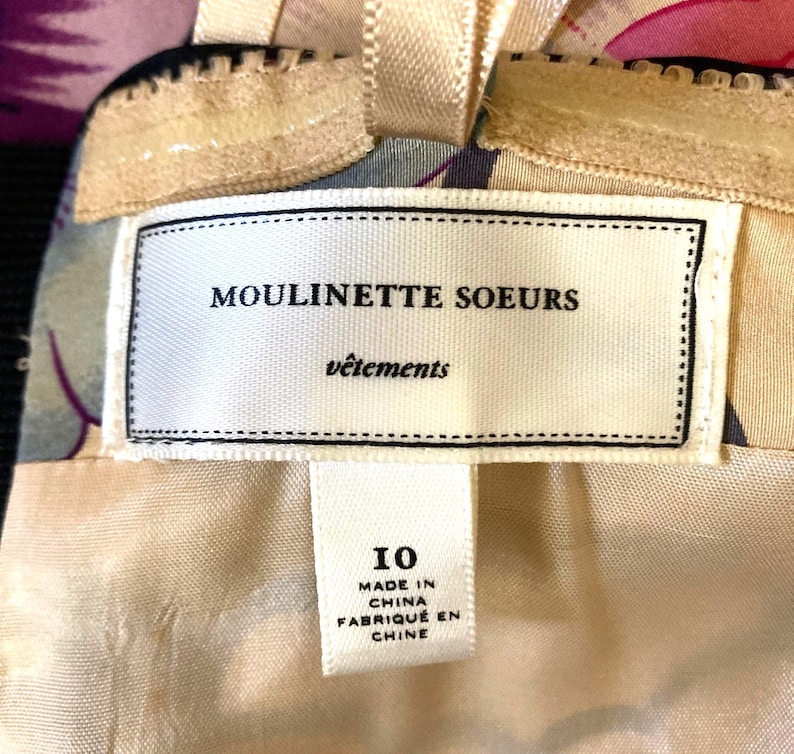 May include: A garment label featuring the text MOULINETTE SOEURS v&ecirc;tements with a size 10 tag below it. The label is white with black text and is attached to a beige fabric with a floral pattern. The tag also states MADE IN CHINA.