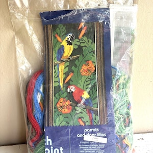 May include: A needlepoint kit in a clear plastic bag featuring a printed design of two parrots and tiger lilies. The label reads Parrots and tiger lilies, Candamar Designs Inc. The kit includes a mesh canvas and bundles of colorful wool yarn.