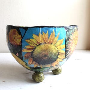 May include: A ceramic planter with a blue, green, and yellow floral design featuring sunflowers. The planter has three legs and is made of paper mache.