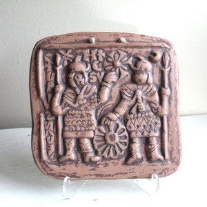 May include: A terracotta wall plaque with a raised relief design of two figures in armor holding weapons. The plaque is brown and has a textured surface.