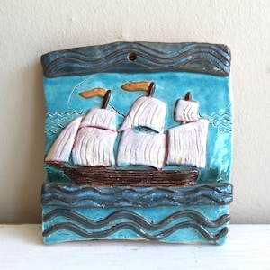 Può includere: Un'applique da parete in ceramica raffigurante una nave a vela. L'opera d'arte ha una forma rettangolare, con uno sfondo blu e una nave marrone con vele bianche. Le onde e il cielo sono blu, con accenti arancioni sulle bandiere.