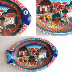 Puede incluir: Un plato de servir de madera con forma de pez colorido con una escena pintada de un pueblo con casas, árboles y personas. El plato está decorado con pintura roja, azul, verde y amarilla.