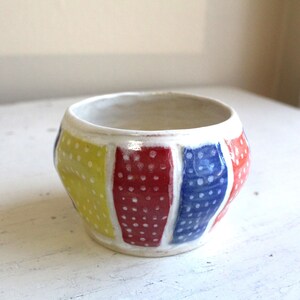 May include: A white ceramic bowl with a rounded shape and a geometric design. The bowl is decorated with yellow, red, and blue polka dots.