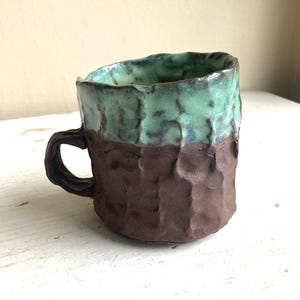 May include: A handmade ceramic mug with a textured surface. The top half is a seafoam green color, and the bottom half is a dark brown. The mug has a small handle and a unique, organic shape, perfect for coffee or tea.