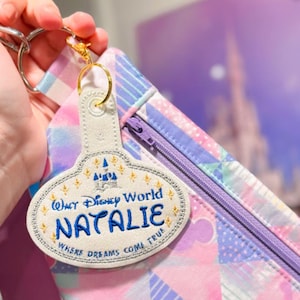 Puede incluir: Un llavero blanco y dorado con el nombre "Natalie" bordado en azul, junto con "Walt Disney World" y "Where Dreams Come True". La etiqueta está sujeta a una bolsa de colores pastel con cremallera.