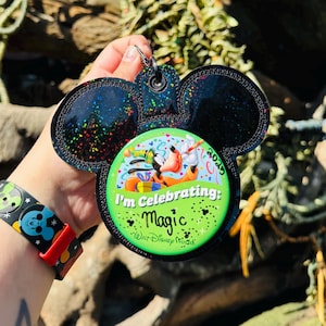 May include: Black Mickey Mouse ear keychain with a green and white button that says "I'm Celebrating: Magic" with Goofy and Pluto. The keychain has a black glitter design and a silver keyring. A Disney MagicBand is also visible.