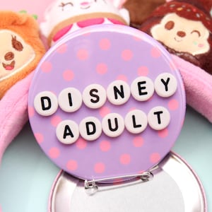 May include: A pink plush headband featuring a circular purple button with pink polka dots and the text DISNEY ADULT in white beads with black letters. The headband is decorated with several small plush character figures in various colors.