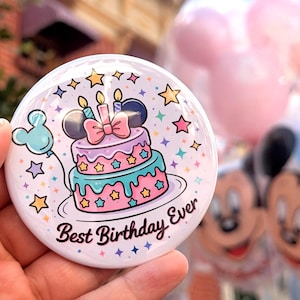 May include: A round white button featuring a colorful illustration of a two-tiered birthday cake with a pink bow, candles, and mouse ears. The design includes small stars and the text Best Birthday Ever in a dark script font on a white background.