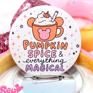 May include: A circular pinback button featuring a white background with a graphic of an orange mug filled with whipped cream and a small pumpkin. The text reads PUMPKIN SPICE & everything MAGICAL in orange, pink, and black lettering.