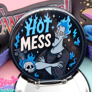 Peut inclure: Un badge rond épinglé illustrant un personnage à la peau bleue avec des cheveux en flammes bleues, les bras croisés et une broche en forme de crâne. Le texte HOT MESS est écrit en lettres blanches et bleues, entouré de flammes et d'un crâne.