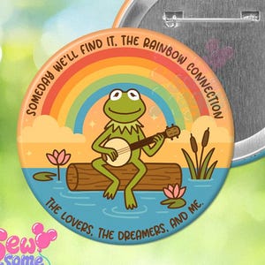May include: A round pinback button featuring a green frog sitting on a log playing a banjo in front of a rainbow. The text reads: SOMEDAY WE'LL FIND IT, THE RAINBOW CONNECTION. THE LOVERS, THE DREAMERS, AND ME. The back shows a metal pin mechanism.