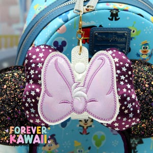 May include: A purple bow-shaped keychain with a light purple embroidered design, attached to a gold-colored clasp. The bow has a white border and is set against a background of a blue backpack with Disney-themed characters and the text "FOREVER KAWAII".