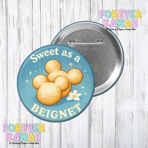 Peut inclure: Un bouton rond avec un fond bleu clair portant l'inscription "Sweet as a Beignet". Le bouton présente une illustration de beignets brun doré et une fleur blanche. Le bouton est un accessoire amusant.