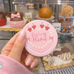 May include: A pink round button with the text "Dishey Mama" in a heart-shaped design, adorned with red cherries and bows. The button is held in front of a display case filled with chocolate-covered apples and treats.