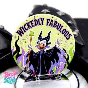 May include: A round pinback button featuring a cartoon duck dressed as a villain with black horns and a purple cape, holding a staff with a green orb. The light yellow background displays the text WICKEDLY FABULOUS in purple with green flames.