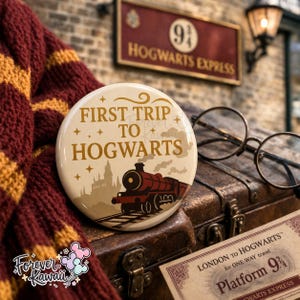 Op de afbeelding: Een ronde, crèmekleurige knoop met de tekst "FIRST TRIP TO HOGWARTS" in gouden letters. De knoop heeft een treinillustratie en sterren. De knoop is geplaatst op een vintage koffer, met een Hogwarts Express-bord op de achtergrond.