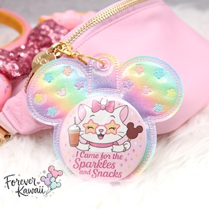 May include: A pastel rainbow-colored keychain in the shape of a mouse head. The keychain features a cartoon cat with sunglasses, holding a drink and a chocolate treat. Text on the keychain reads "I Came for the Sparkles and Snacks."