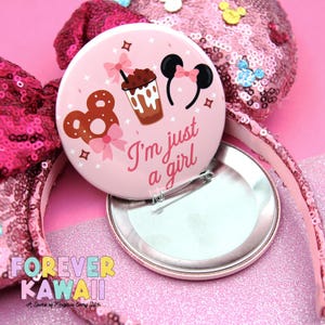 May include: A pink button with the text "I'm just a girl" and illustrations of a pretzel, drink, and mouse ears. The button is set against a pink sequined background and a pink headband.