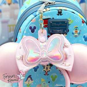 May include: A pink Minnie Mouse bow-shaped keychain with iridescent accents hangs in front of a light blue Disney Parks Loungefly backpack. The backpack features a pattern of Disney characters and icons. The keychain is attached with a gold-colored clasp.