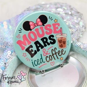 May include: A round, teal button with the words "MOUSE EARS iced & coffee" in pink, black, and brown. The button features a Minnie Mouse bow and a coffee drink illustration. The button is attached to a silver pin back.