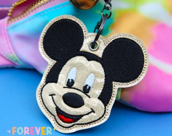 Könnte beinhalten: Ein Mickey Mouse-förmiger Schlüsselanhänger mit goldenem und schwarzem Design. Der Schlüsselanhänger ist an einer bunten Tasche mit goldenem Reißverschluss befestigt. Der Text "Forever Kawaii" befindet sich am unteren Rand des Bildes.