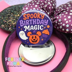 May include: A purple button with the words "Spooky Birthday Magic" and Halloween-themed graphics, including a ghost, pumpkin, and cupcake. The button is on a pink surface with a black headband and purple bow.