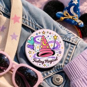 Könnte beinhalten: Ein runder, hellrosa Button mit einem Zaubererhut, Sternen und dem Text "Feeling Magical". Der Hut ist rosa mit gelben Sternen und einem Mond. Ein Zauberstab und Luftballons sind ebenfalls abgebildet. Der Button befindet sich auf einer Jeansjacke.