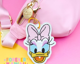 May include: A pink fanny pack with a gold zipper and hardware, featuring a Daisy Duck keychain. The keychain is an embroidered patch with a pink bow, blue eyes, and a yellow beak. The text "FOREVER KAWAII" is visible.