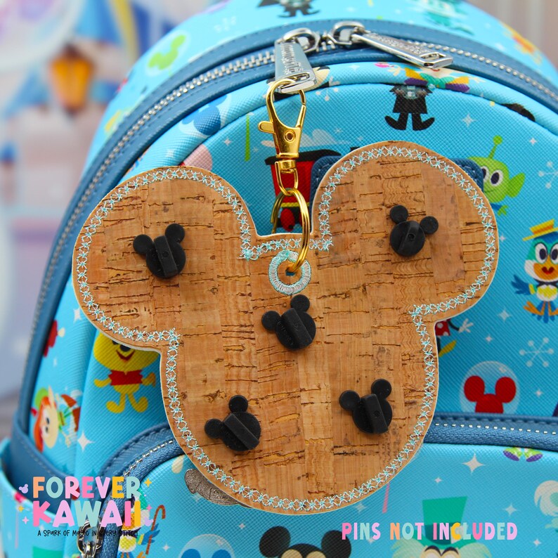 May include: A Mickey Mouse-shaped cork keychain with black Mickey head pins. The keychain has a gold clasp and a light blue stitched border. The background is a blue backpack with cartoon characters.