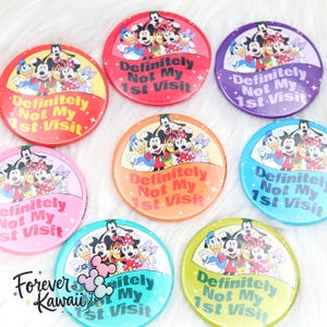May include: A selection of circular badges in various colours, each displaying Disney characters including Mickey and Minnie Mouse, Donald Duck, and Goofy. The text "Definitely Not My 1st Visit" is featured on each badge.
