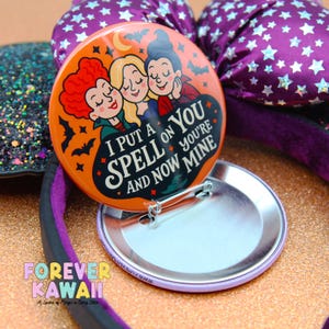 May include: An orange button with a cartoon image of three witches and the text "I PUT A SPELL ON YOU AND NOW YOU'RE MINE." The button is next to a purple bow with white stars.