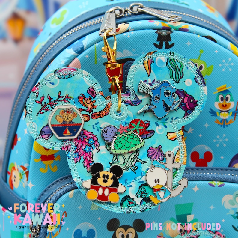 May include: A light blue backpack with a Mickey Mouse ear-shaped charm featuring various Disney character pins. The charm is attached with a gold-colored clasp. The text "FOREVER KAWAII" is visible on the backpack.