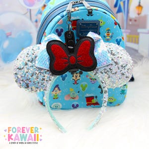 May include: A light blue sequined Minnie Mouse ear headband with a red bow and a matching blue backpack. The backpack features Disney character graphics. A black Minnie Mouse bow keychain hangs from the backpack zipper.