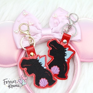 Op de afbeelding: Twee sleutelhangers met een zwart silhouet, blauwe en paarse details en rode leren bandjes. Ze zijn geplaatst tegen een roze Minnie Mouse oren haarband met een met parels versierde strik. De sleutelhangers hebben gouden en zilveren sluitingen.