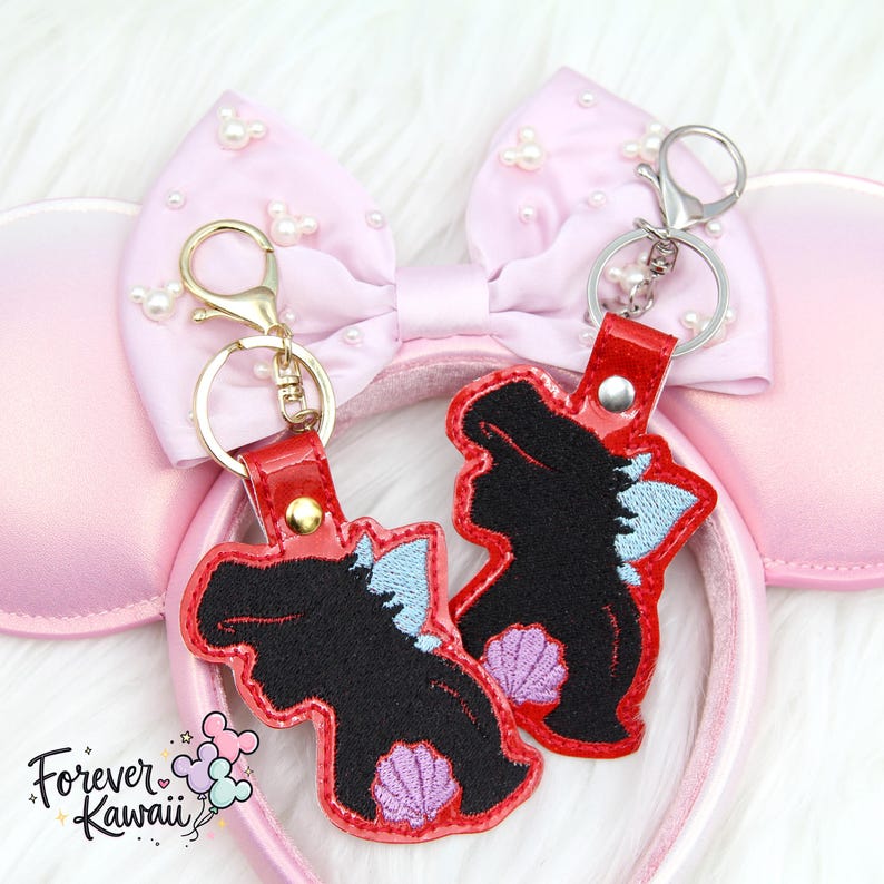 Op de afbeelding: Twee sleutelhangers met een zwart silhouet, blauwe en paarse details en rode leren bandjes. Ze zijn geplaatst tegen een roze Minnie Mouse oren haarband met een met parels versierde strik. De sleutelhangers hebben gouden en zilveren sluitingen.