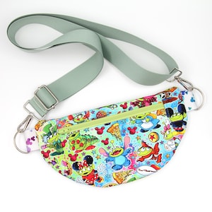 May include: A green, blue, and white fanny pack with a zipper closure and a green adjustable strap. The fanny pack features a variety of cartoon characters, including aliens, a caterpillar, and Mickey Mouse.