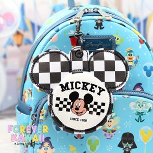 May include: A Mickey Mouse-shaped keyring with a black and white chequered pattern and a circular emblem featuring Mickey's face and the word "MICKEY" above the text "SINCE 1928". The keyring is attached to a light blue backpack.