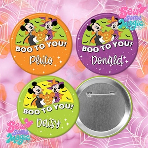 Op de afbeelding: Drie ronde buttons in oranje, paars en limoengroen, elk met Mickey en Minnie Mouse in Halloween-kostuums die een pompoen vasthouden. De tekst luidt BOO TO YOU! met de namen Pluto, Donald of Daisy. Eén achterkant van een button is zichtbaar.