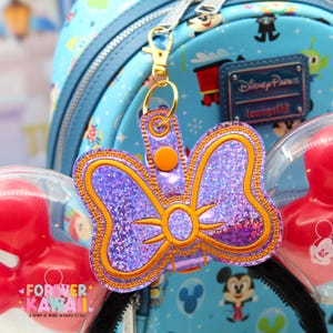May include: A purple glitter bow-shaped keychain with gold trim and a gold clasp. The keychain is in front of a light blue Disney-themed backpack and red Mickey Mouse ears.