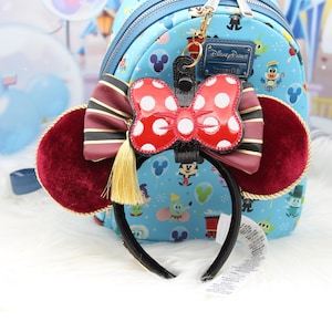 May include: A Minnie Mouse ear headband with a red polka dot bow and burgundy velvet ears. The headband has a black band and a gold tassel. A light blue backpack with Disney characters is in the background.