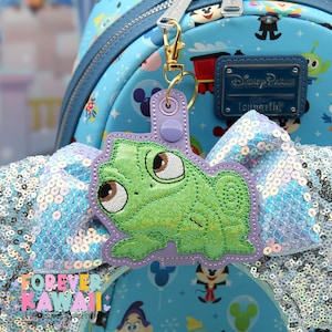 May include: A green Pascal the chameleon keychain with glitter accents, attached to a gold-colored clasp. The keychain is set against a light blue Disney Parks Loungefly backpack with a sequined bow.