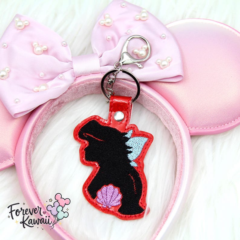 Op de afbeelding: Een roze Minnie Mouse oren haarband met een bijpassende strik versierd met parels. Een rood omrande sleutelhanger met een zwart silhouet van een zeemeermin met een schelp en blauwe accenten hangt aan de strik. De tekst "Forever Kawaii" staat linksonder.