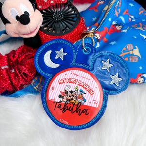 May include: A blue Mickey Mouse-shaped keyring with a red and white striped circle featuring Mickey, Minnie, Donald, and Goofy. The text "My First Launch" and "Tabitha" are visible. Other Disney-themed items are in the background.
