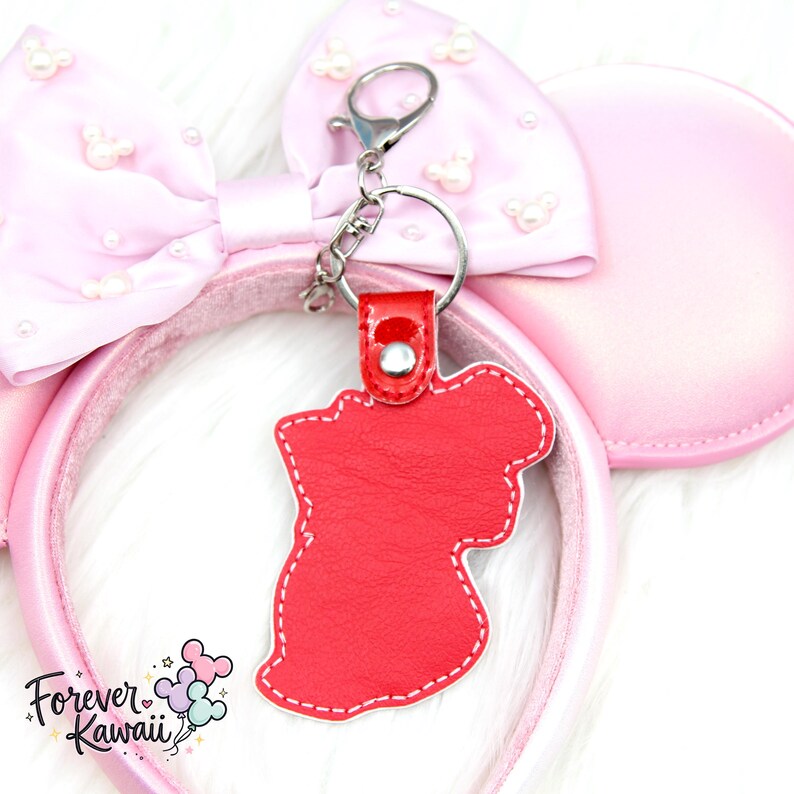 Op de afbeelding: Een rode sleutelhanger van imitatieleer in de vorm van een personage, met witte stiksels en een zilveren sluiting. De sleutelhanger is bevestigd aan een roze haarband met een strik en parelaccenten. De tekst "Forever Kawaii" is zichtbaar.